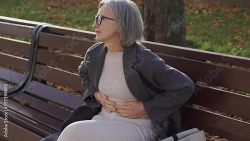 Elegant senior woman frowning in pain sitting on bench in park, feeling acute abdominal pain, suffering gastritis or duodenal ulcer, risk of ruptured appendix or congestion in the gallbladder photo