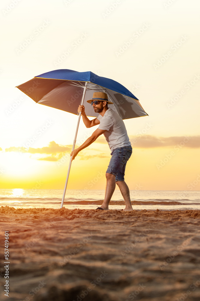 Man in a hat, sunglasses, and t-shirt sticking a large umbrella into the sand on the beach during a summer sunrise