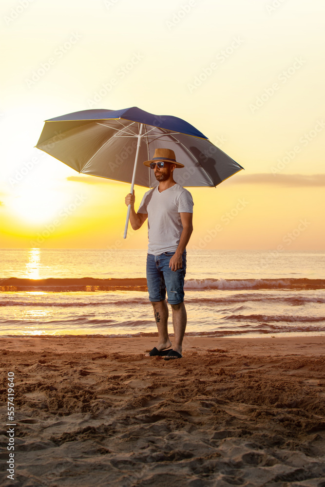 Man in a hat, sunglasses, t-shirt and flip flops standing on the sand at the beach holding a large umbrella during a summer sunrise Man in a hat, sunglasses, t-shirt and flip flops standing on the sand at the beach holding a large umbrella during a summer sunrise