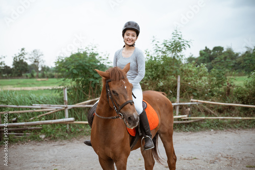 Female equestrian smiling while riding horse and holding reins in outdoor background