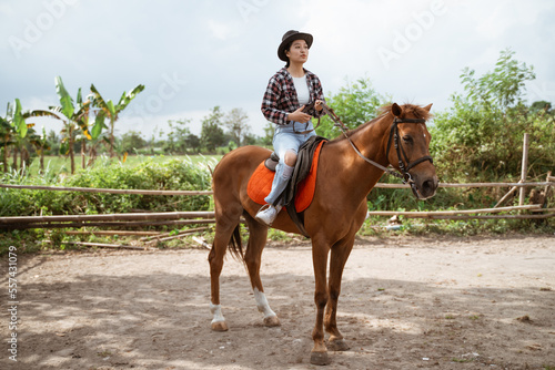 asian woman in cowboy hat holding reins while sitting on horse at ranch