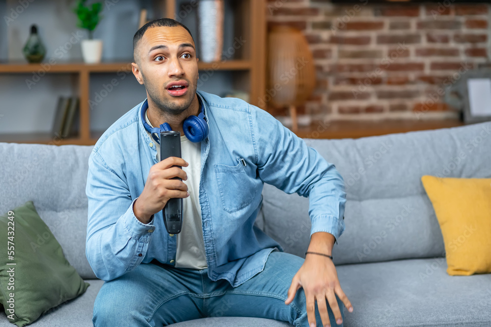 Surprised male sitting on couch at home watching exciting TV show online shocked with cliffhanger ending of detective series, keeping his mouth open Surprised male sitting on couch at home watching exciting TV show online shocked with cliffhanger ending of detective series, keeping his mouth open