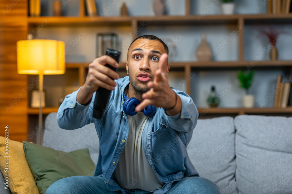 Closeup portrait, young man, sitting on gray couch, watching TV, holding remote, surprised at what he sees, raising his hand. Closeup portrait, young man, sitting on gray couch, watching TV, holding remote, surprised at what he sees, raising his hand.
