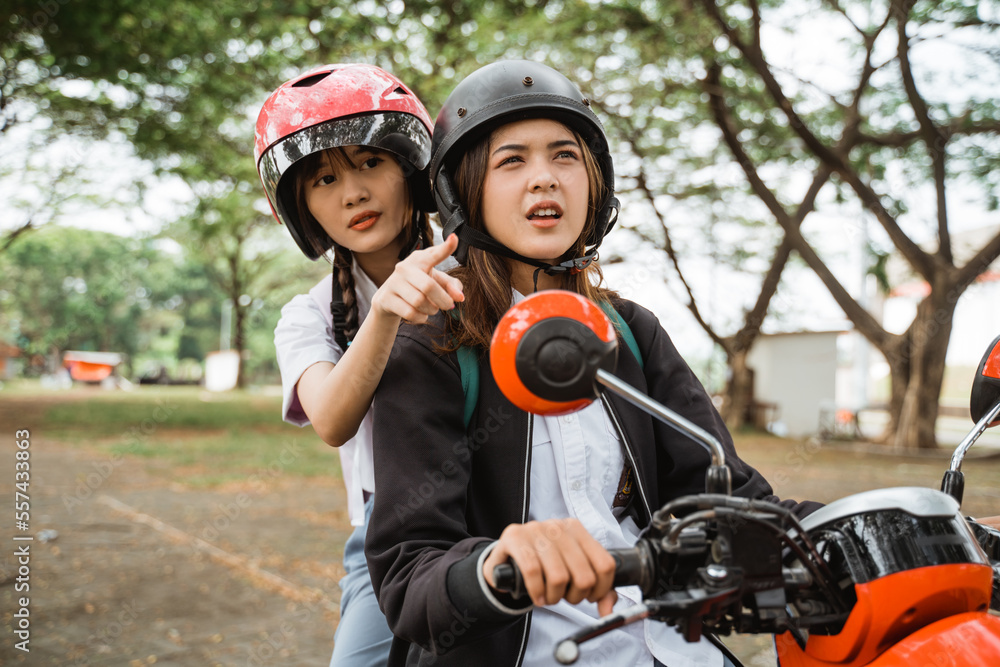 Two high school girls wearing helmets and jackets looking for an address while riding a motorbike