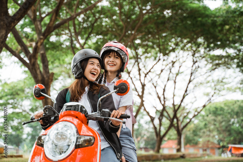 Two student girls wearing helmets and jackets chatting while riding motorbikes together