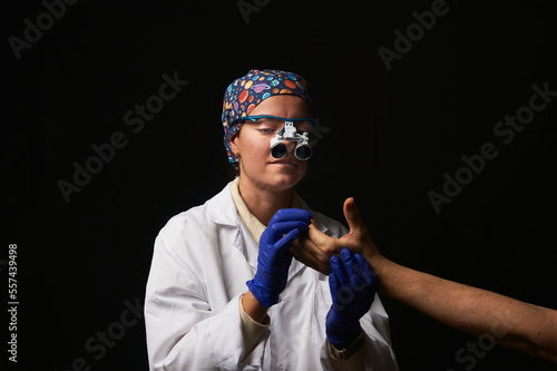 Female doctor in a white coat, blue nitrile gloves, microsurgical goggles, and a headcap examining a patient's hand photo