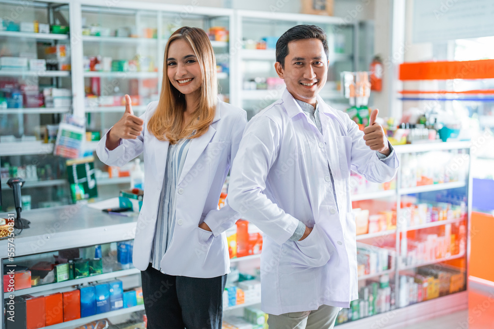 Two smiling asian female and male pharmacists with thumbs up in drugstore Two smiling asian female and male pharmacists with thumbs up in drugstore