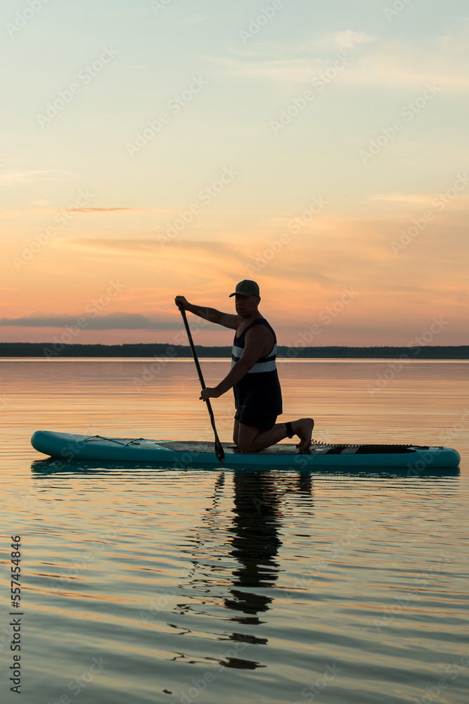 A man in shorts on his knees on a SUP board with an oar against the backdrop of a sunset sky swims in the lake in the evening.