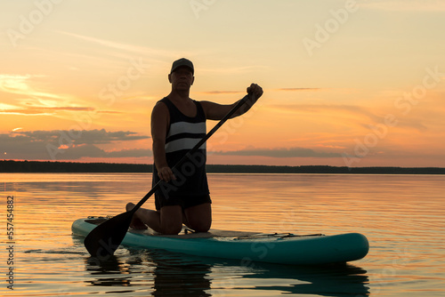 A man in shorts on his knees on a SUP board with a paddle at sunset swims in the water of the lake in the glare from the setting sun.