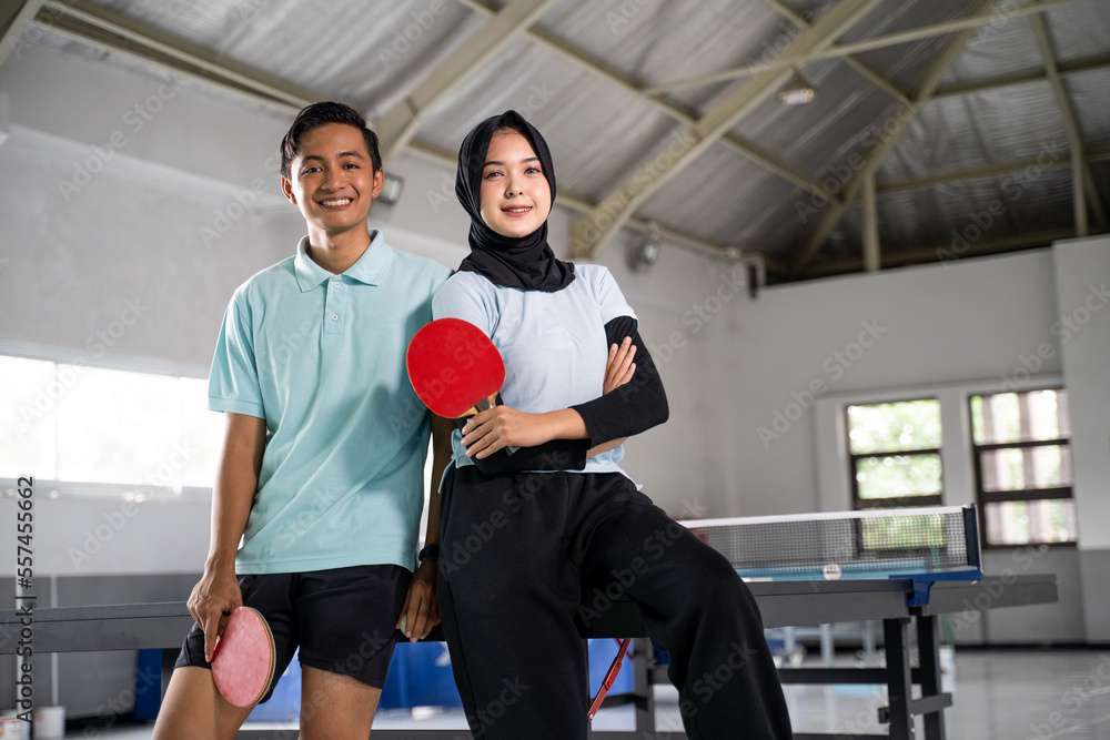 man and woman athlete in hijab standing holding bat with crossed hands on ping pong table background