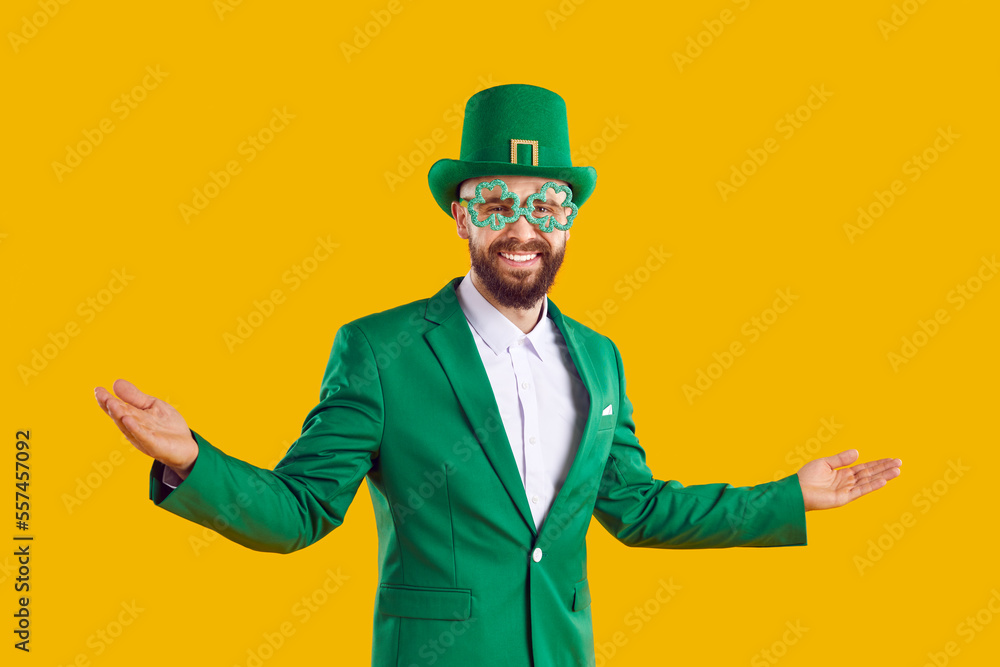 Welcome to St Patrick's Day Party. Happy positive joyful cheerful bearded man wearing green suit, leprechaun hat and funny clover glasses smiling and spreading his hands on yellow studio background