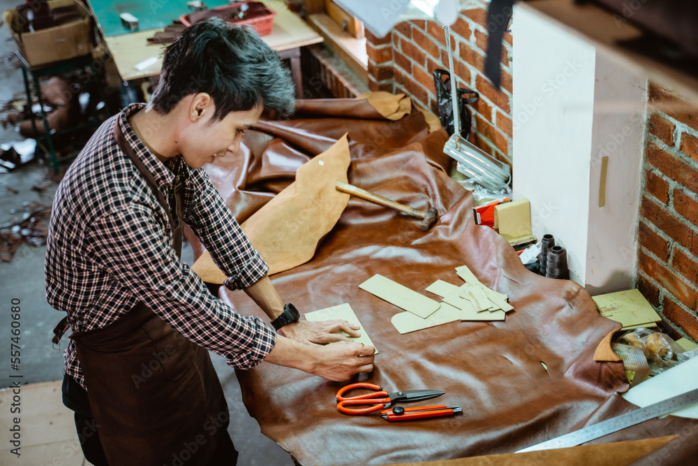 Asian craftsman in apron working with leather clinging pattern paper in workshop Asian craftsman in apron working with leather clinging pattern paper in workshop