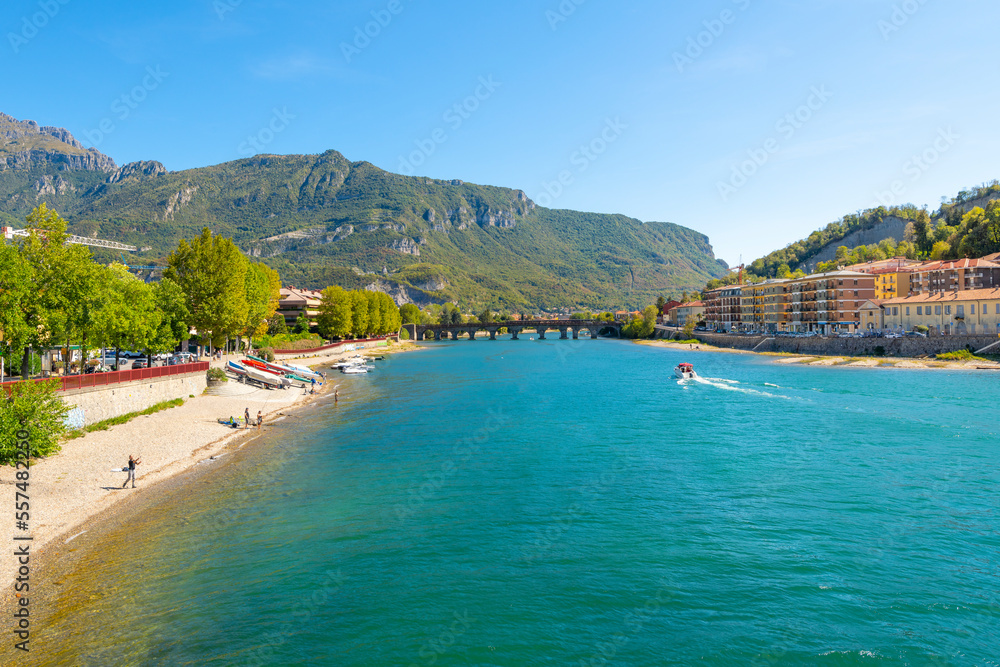 Italians fish from the riverbank of Lake Como and the Adda river near the historic Old Town of Lecco, Italy, with the Ponte Vecchio bridge and Monte Resegone in view.