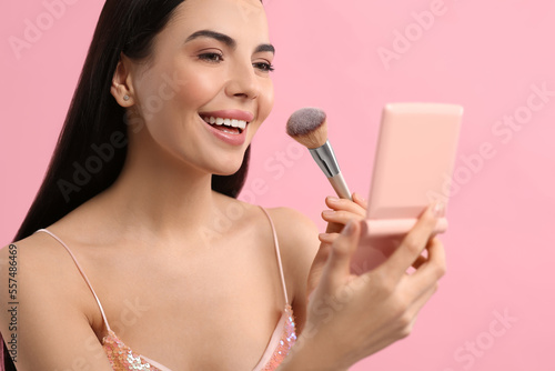 Beautiful woman with cosmetic pocket mirror applying makeup on pink background
