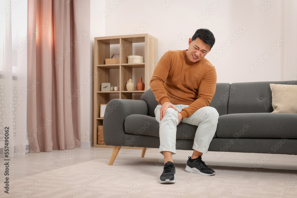 Asian man suffering from knee pain on sofa indoors Asian man suffering from knee pain on sofa indoors