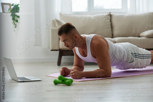 Man sports, watching tape of workout on phone and repeating exercises sports blogger with laptop training online, pumped up man fitness trainer works out at home, the concept of health and body beauty