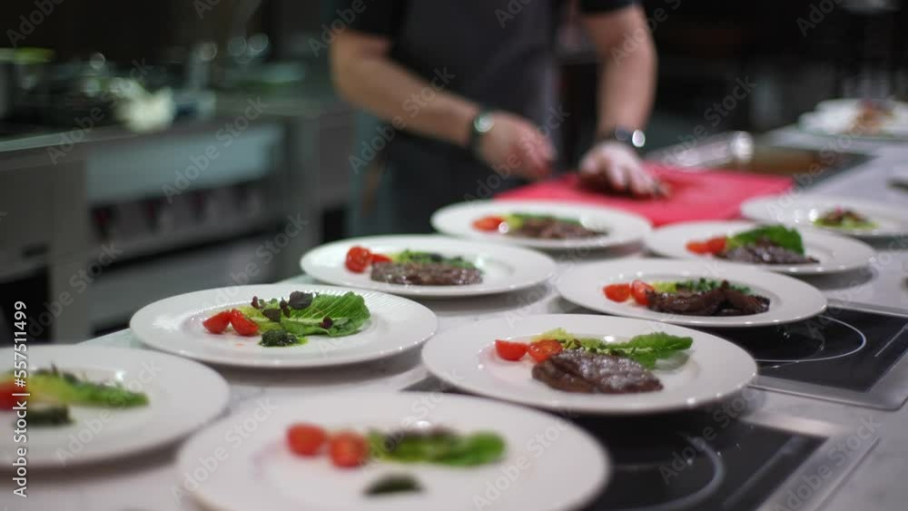 Cropped shot of unrecognizable male chef in gloves cutting grilled or fried meat with kitchen knife and arranges it on plates. Chef preparing delicious dish in restaurant. Shooting in slow motion. Cropped shot of unrecognizable male chef in gloves cutting grilled or fried meat with kitchen knife and arranges it on plates. Chef preparing delicious dish in restaurant. Shooting in slow motion.