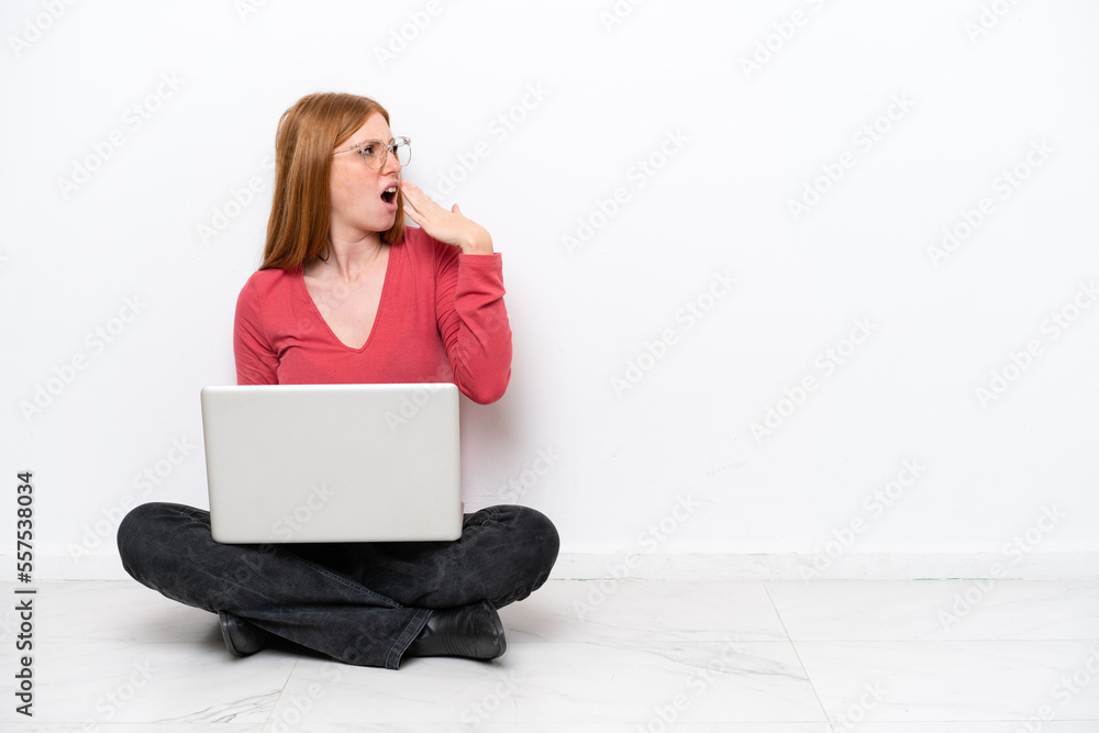 Young redhead woman with a laptop sitting on the floor isolated on white background yawning and covering wide open mouth with hand Young redhead woman with a laptop sitting on the floor isolated on white background yawning and covering wide open mouth with hand
