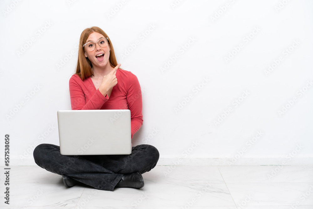 Young redhead woman with a laptop sitting on the floor isolated on white background surprised and pointing side Young redhead woman with a laptop sitting on the floor isolated on white background surprised and pointing side