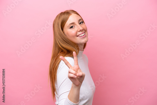 Young redhead woman isolated on pink background smiling and showing victory sign