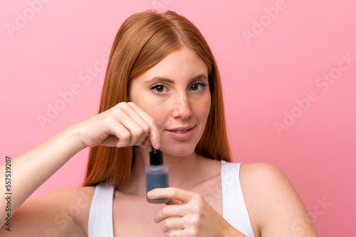 Young redhead woman isolated on pink background holding a serum. Close up portrait