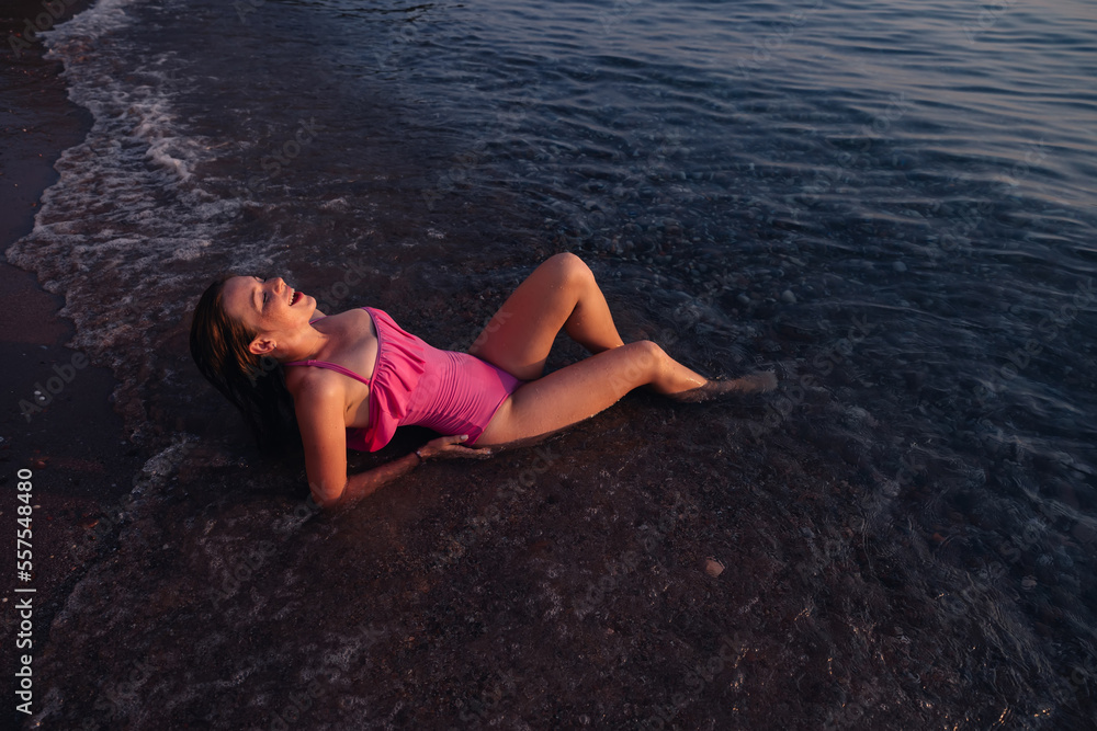 Happy woman in pink swimsuit laying on a beach in the water with closed eyes at the sea.