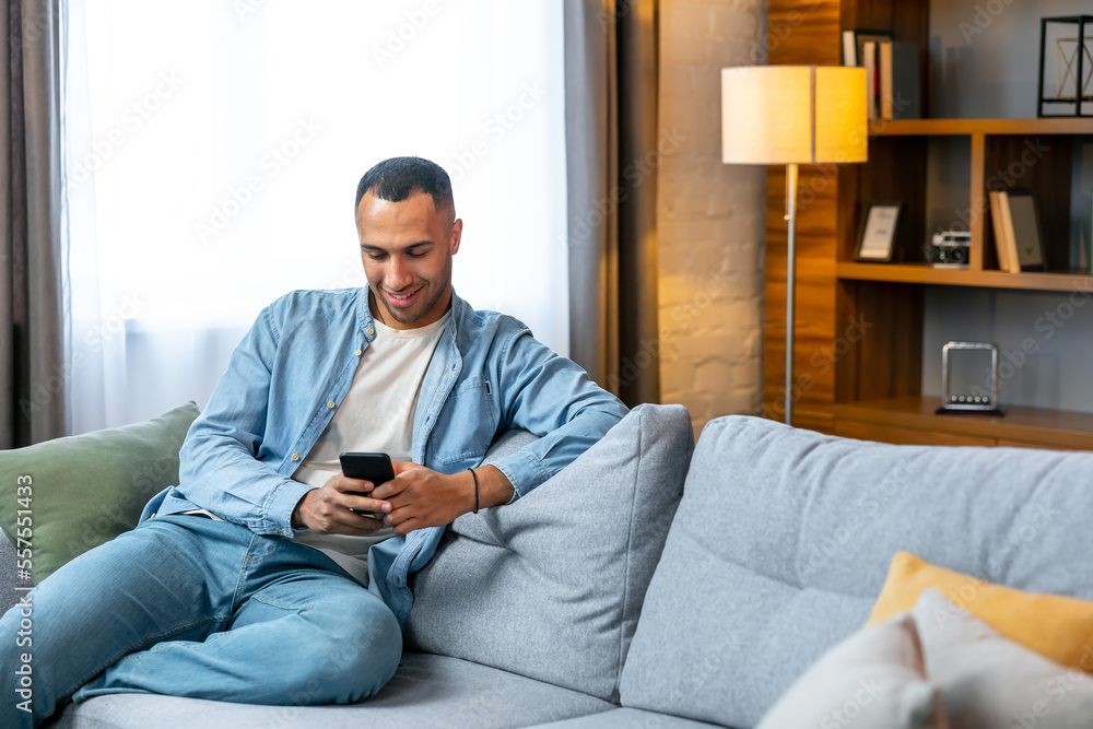 Portrait of an attractive smiling young african man wearing casual clothes sitting on a couch at the living room, using mobile phone