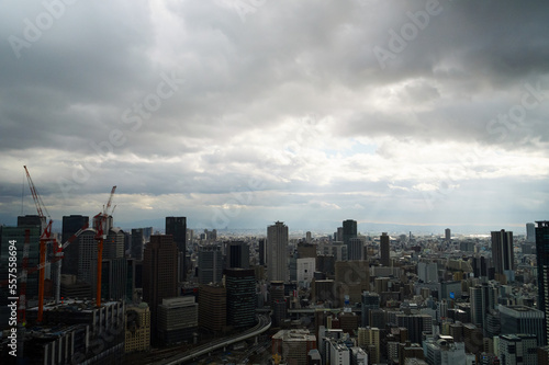 Overhead view of Osaka's Umeda area from a hill on a cloudy day, sunlight shining through a gap in the clouds. © 隼人 岩崎