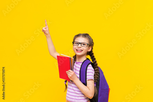 A little joyful girl holds notebooks in her hand and points to the side at your advertisement.