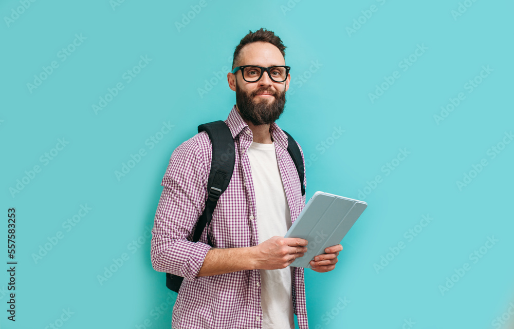 Portrait of handsome smiling student in casual wear with backpack while holding digital tablet over blue studio background Portrait of handsome smiling student in casual wear with backpack while holding digital tablet over blue studio background