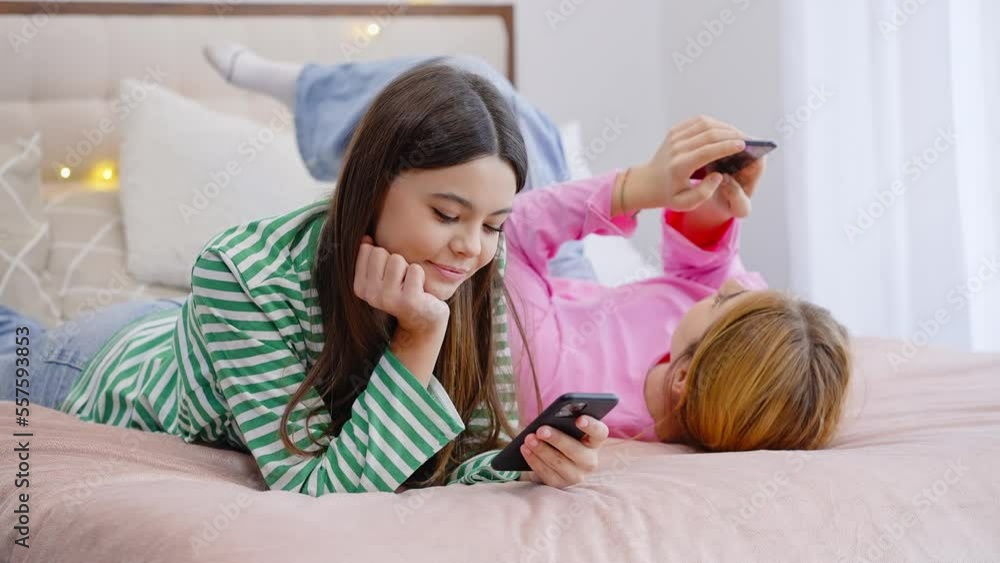 Relaxed two charming adolescent friends girls lying on the bed and scrolling news feed on smartphone Relaxed two charming adolescent friends girls lying on the bed and scrolling news feed on smartphone