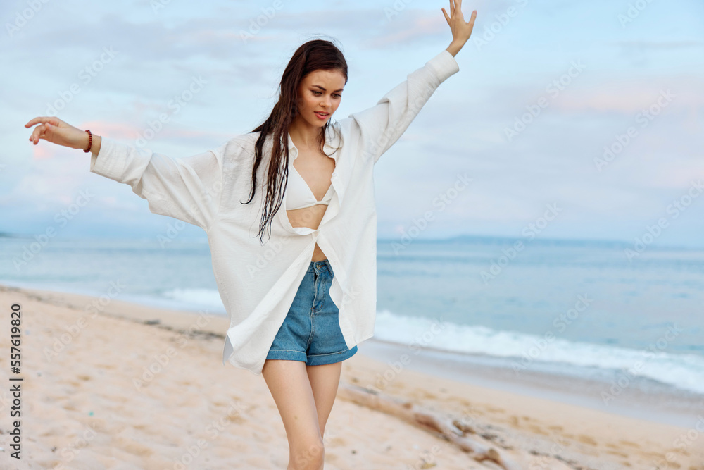 Happy tanned woman in white swimsuit shirt and denim shorts walks on the beach on the sand by the ocean with wet hair after swimming, sunset light and pink clouds in Bali Happy tanned woman in white swimsuit shirt and denim shorts walks on the beach on the sand by the ocean with wet hair after swimming, sunset light and pink clouds in Bali