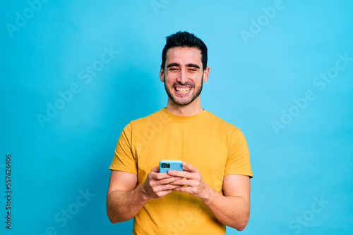 Young bearded male in casual clothes browsing mobile phone while standing against blue background