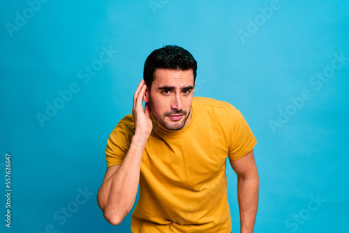 Young bearded male in yellow shirt touching the ear listening while standing against blue background