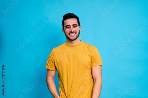 Positive young bearded male in yellow shirt smiling and looking at camera while standing against blue background © The Attico Studio