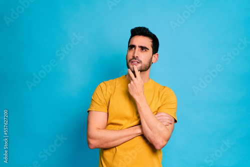 Young bearded male in yellow shirt touching chin and looking at camera while standing against blue background