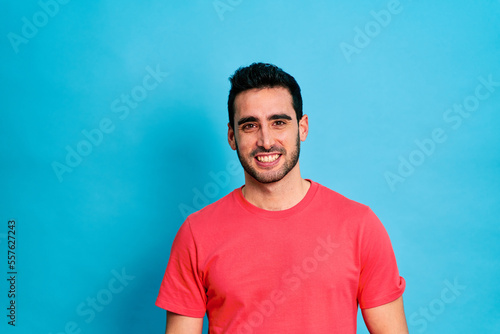 Positive young bearded male in red shirt smiling and looking at camera while standing against blue background