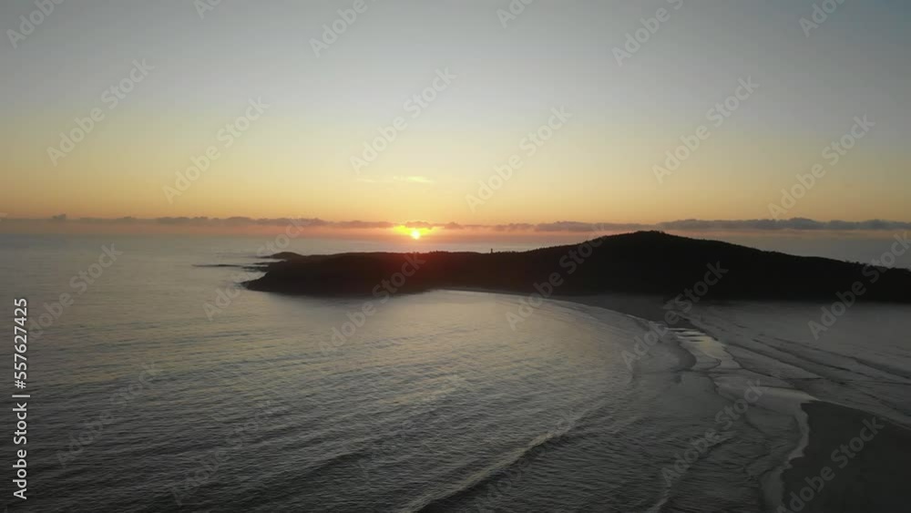 Drone video flying toward Shark Island, Final Bay, as the sun rises behind it. Port Stephens lighthouse silhouetted against the bright sky.