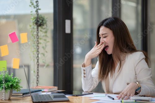 Portrait of a tired sleepy businesswoman yawning, working at a desk in the office in front of a computer. Bored young female freelancer working on PC at home. Overwork and sleep deprivation concept. © Songsak C