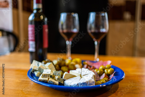 A plate filled with different kinds of cheese  olives  peppers  sausage  garlic and cucumber and a bottle of red wine with two glasses.