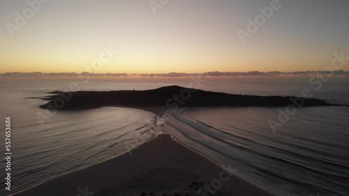 Drone video flying toward Shark Island, Final Bay, as the sun rises behind it. Port Stephens lighthouse silhouetted against the bright sky.
