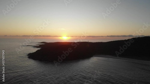 Drone video flying toward Shark Island, Final Bay, as the sun rises behind it. Port Stephens lighthouse silhouetted against the bright sky.