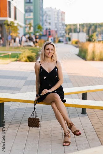Cheerful blonde woman in a black dress with a handbag and sunglasses is resting on a bench in the city in summer © Sunshine