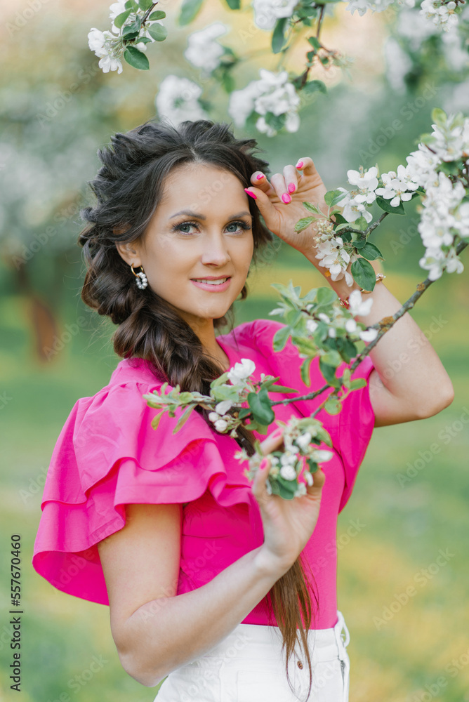 Fototapeta premium A beautiful young European woman with beautiful makeup and braids in spring in the park, in a blooming apple orchard