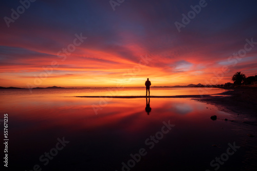 Idyllic orange sunrise on the beach of Los Urrutias, in the Mar Menor, Cartagena, Region of Murcia with the clouds reflecting in the calm waters of the sea and a silhouette of a man watching the sunri