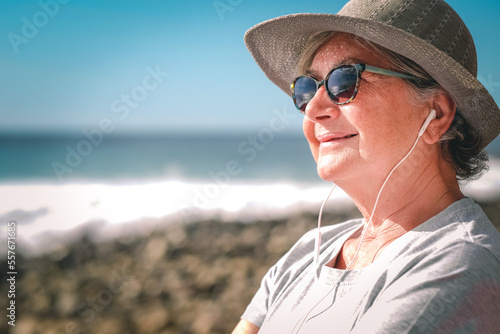 Portrait of beautiful senior woman listening music by earphones - elderly happy lady on the beach enjoying free time and retirement. Horizon over sea #557671685