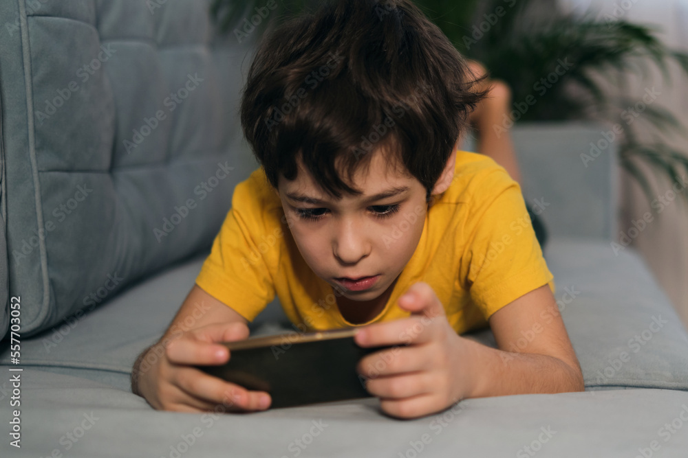 boy with smartphone or game console in hands, playing or engaging over social media while lying on a sofa. kids and gadgets concept