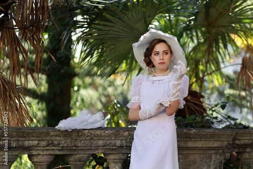 Fotografie Young woman in white edwardian style dress and white big hat standing in old abandoned castle