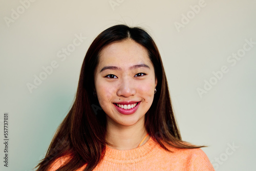 Cheerful Asian female in pink sweater smiling and looking at camera while standing on cream background in studio