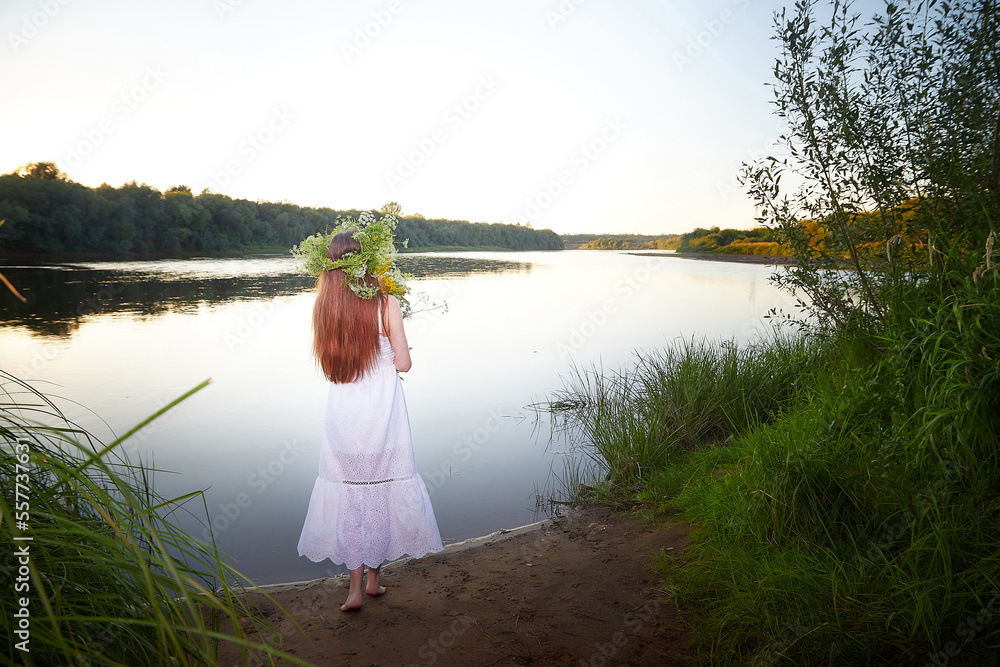 Fototapeta premium Portrait of beautiful slavic girl with long hair with flower crown near water of river or lake on nature in sunny evening. National selebration of summer holiday of Ivan Kupala